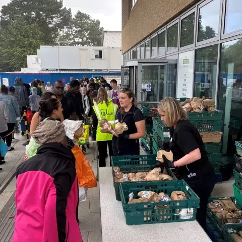 Volunteers hand out bread and fresh produce from crates to a queue of people.