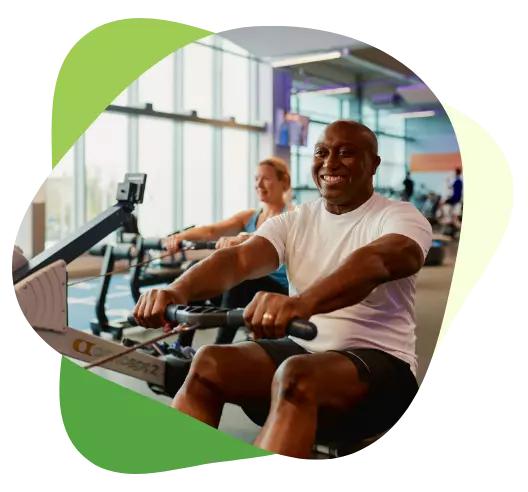 Smiling man using a rowing machine at the gym, with others exercising in the background.