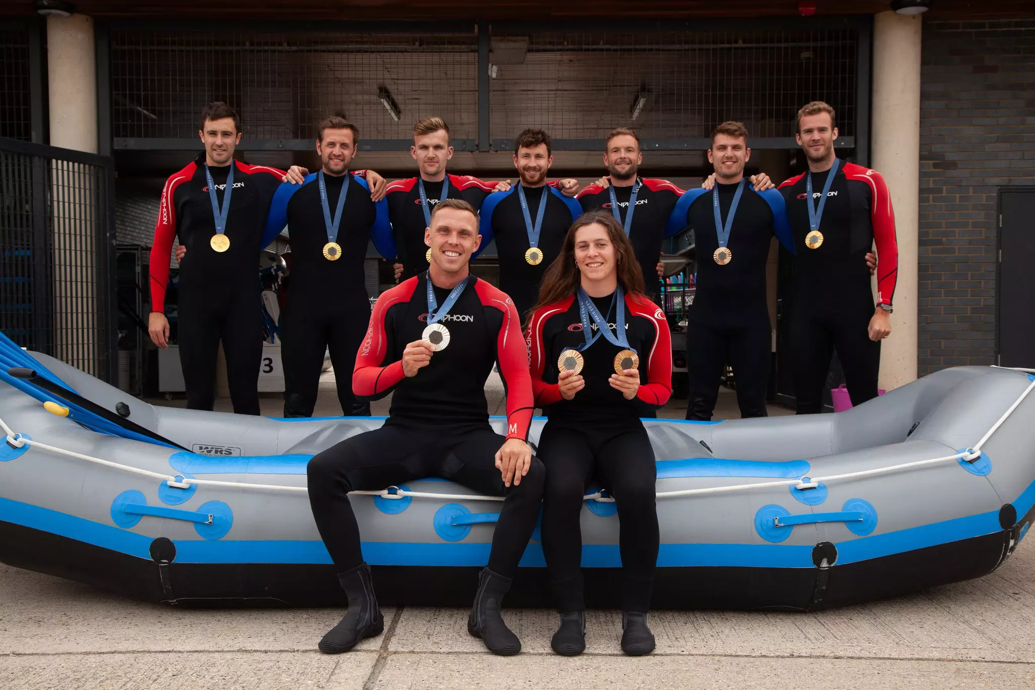 Team of athletes in wetsuits posing with medals in front of a raft, two seated and smiling.