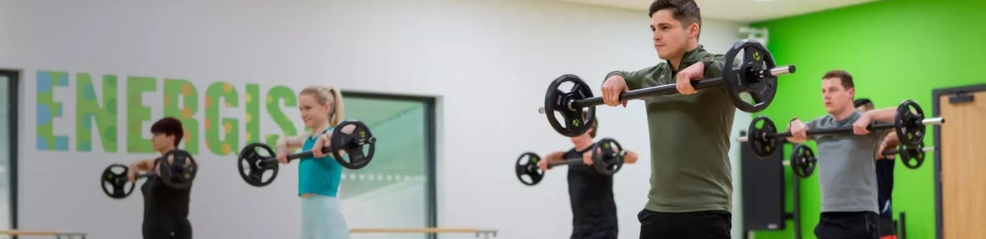 Group of people lifting barbells in a bright gym with green accent walls.