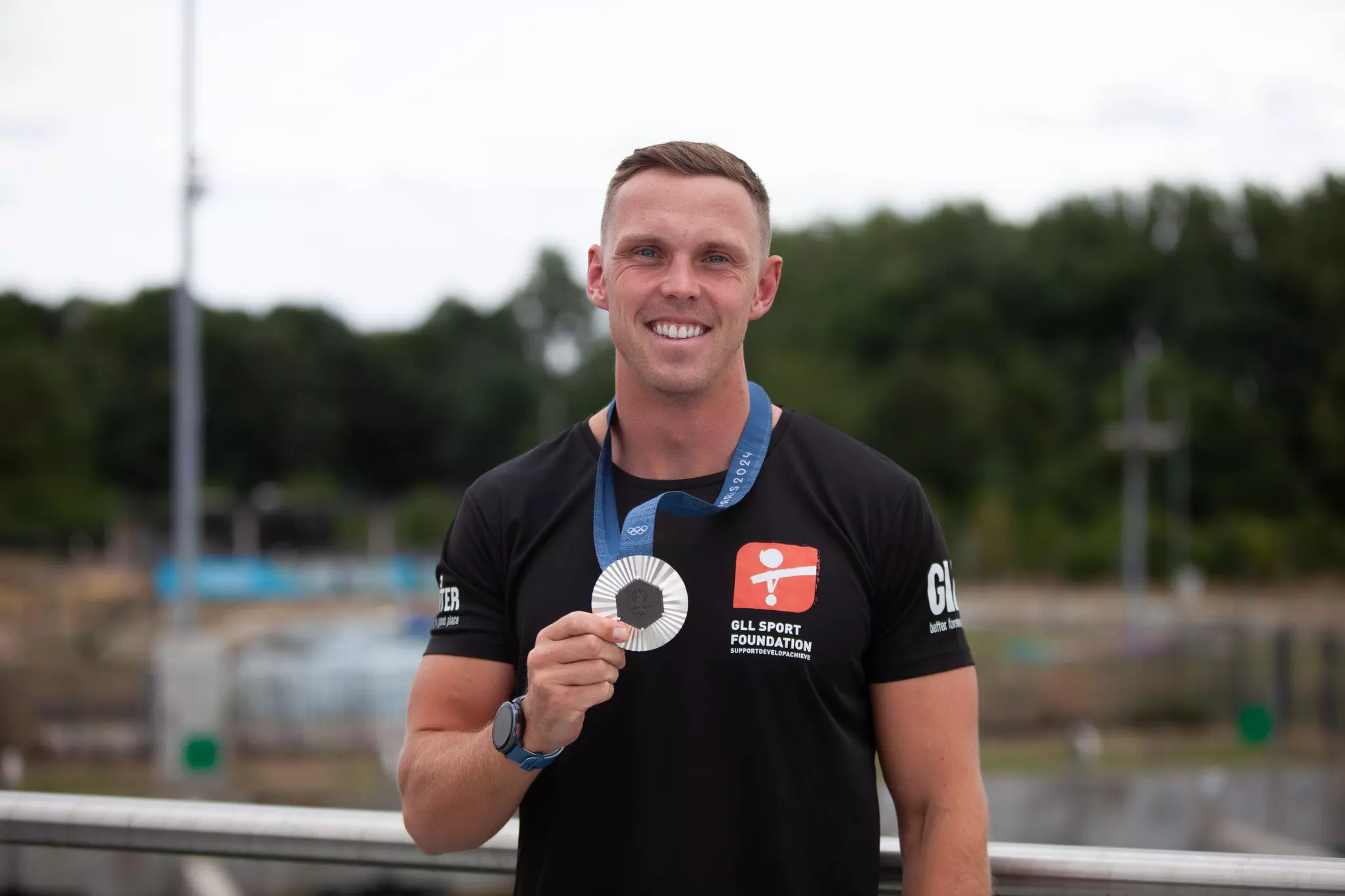 Man in black shirt holding a medal by water.