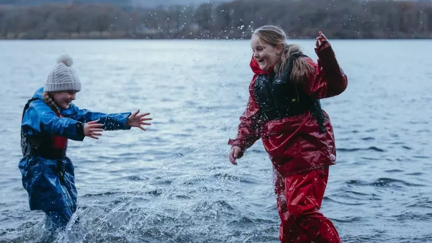 Two children in waterproof gear laughing and splashing in a lake.