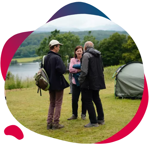 Three adults talking outdoors near a tent with a lake in the background.