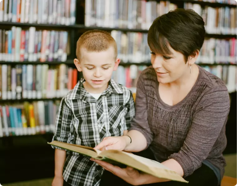 Adult reading a book with a young boy in a library, surrounded by shelves of books.