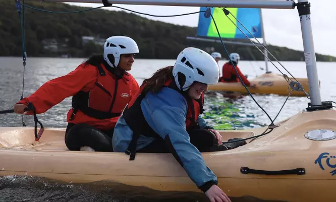 Two young people in helmets and life jackets sailing on a small boat.