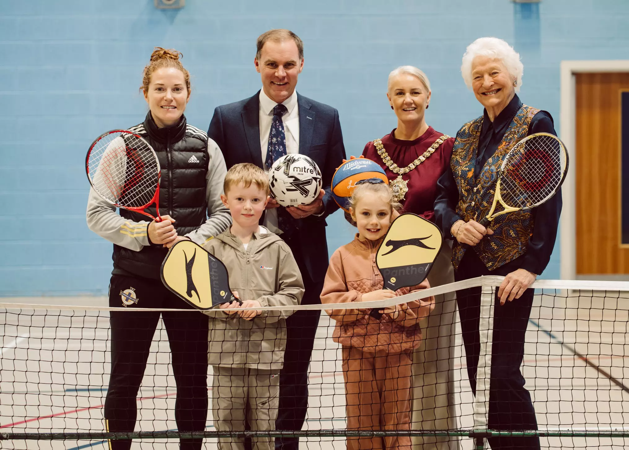 Group of adults and children standing on an indoor court holding sports equipment.