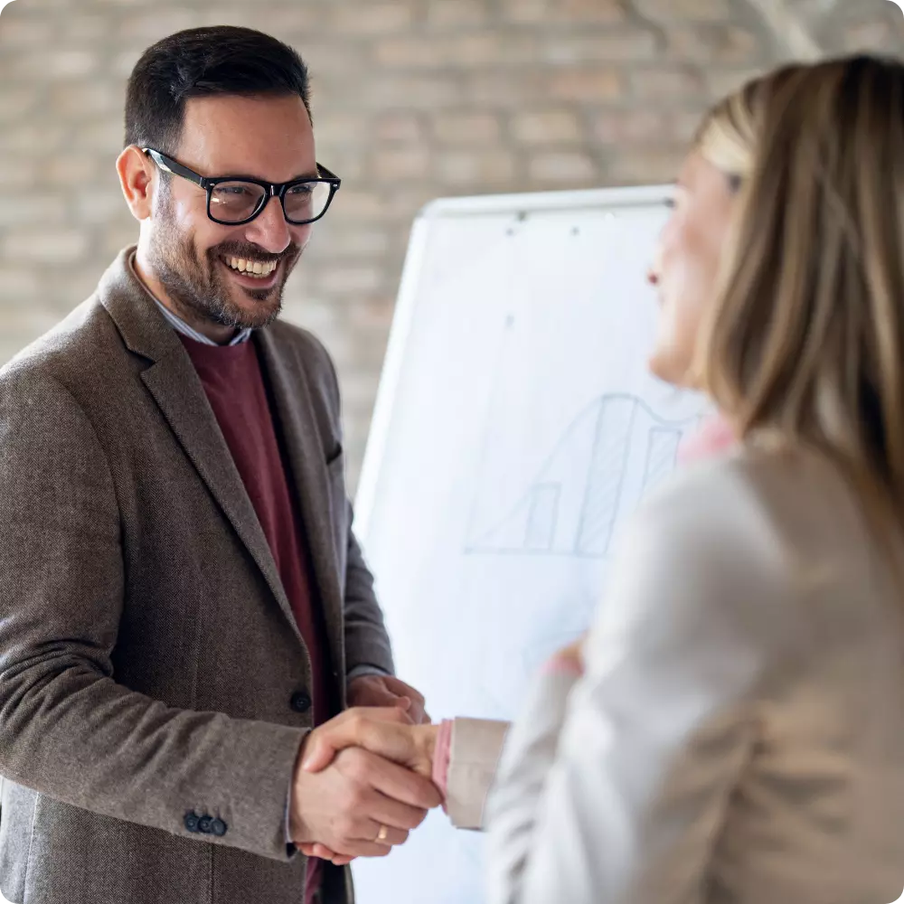 Man in glasses smiles and shakes hands with a woman in front of a flipchart showing a graph.