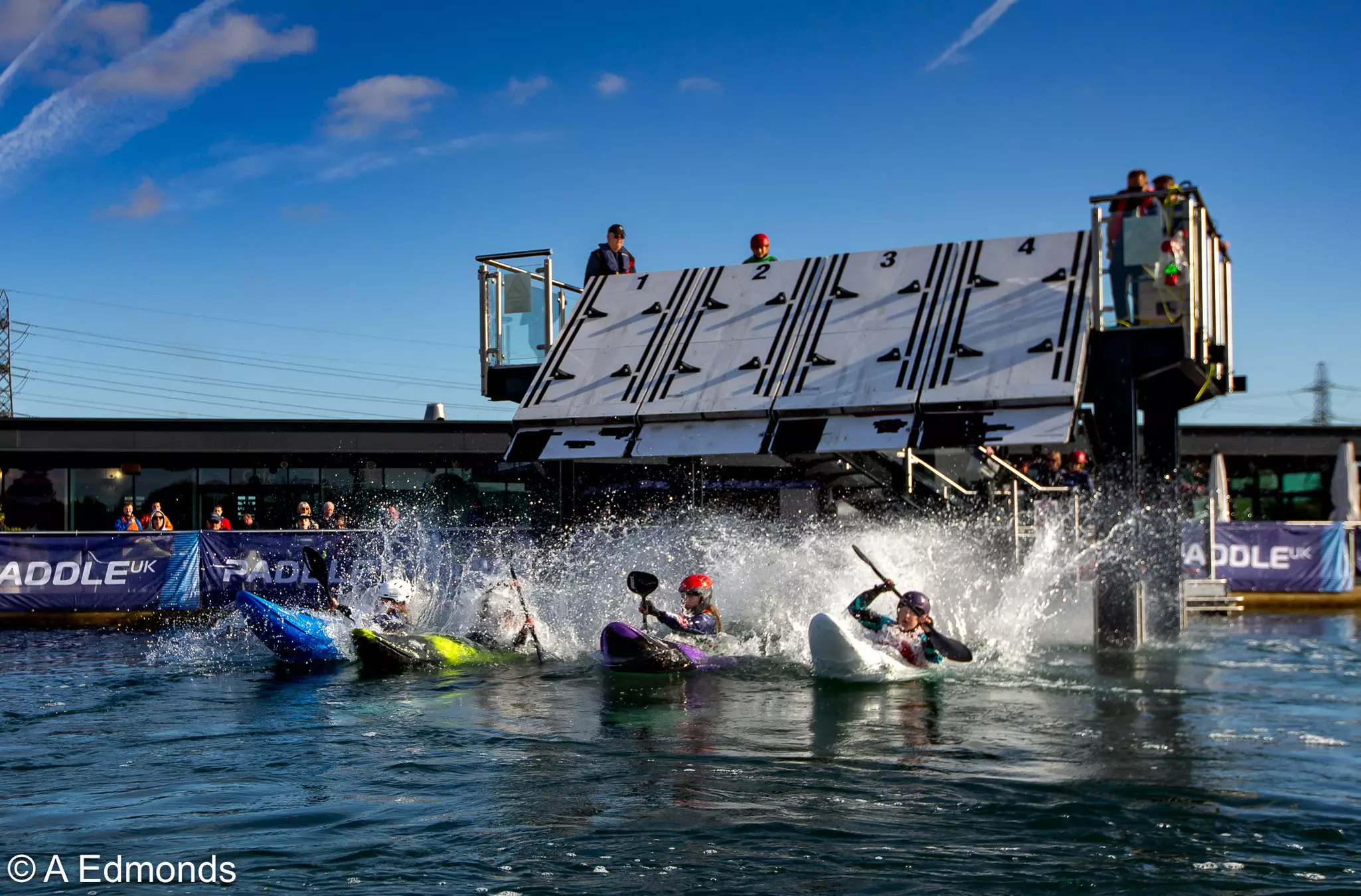 Kayakers racing from a start gate, splashing into the water at a competitive Paddle UK event.