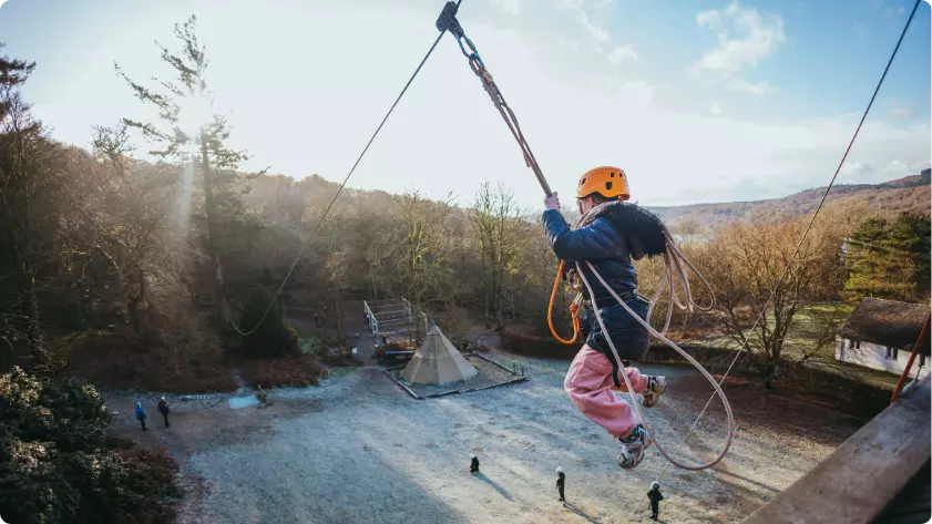 Child in a helmet gliding on a high ropes zip wire above a forest clearing.