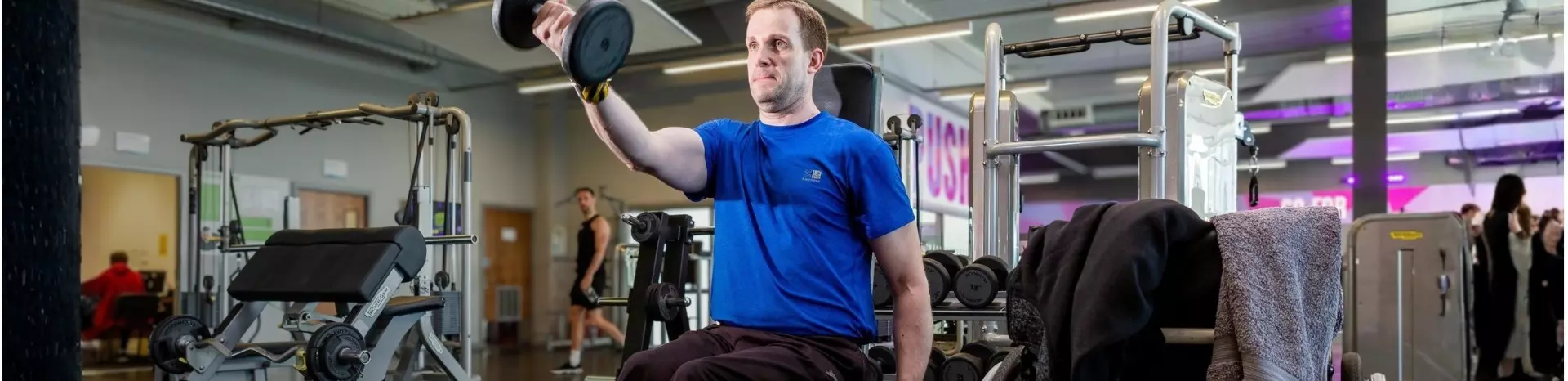 Person in wheelchair lifting dumbbell in a gym with other members in background. 
