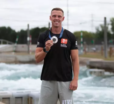 Athlete smiling and holding a medal at a water sports venue, wearing GLL Sport Foundation kit.