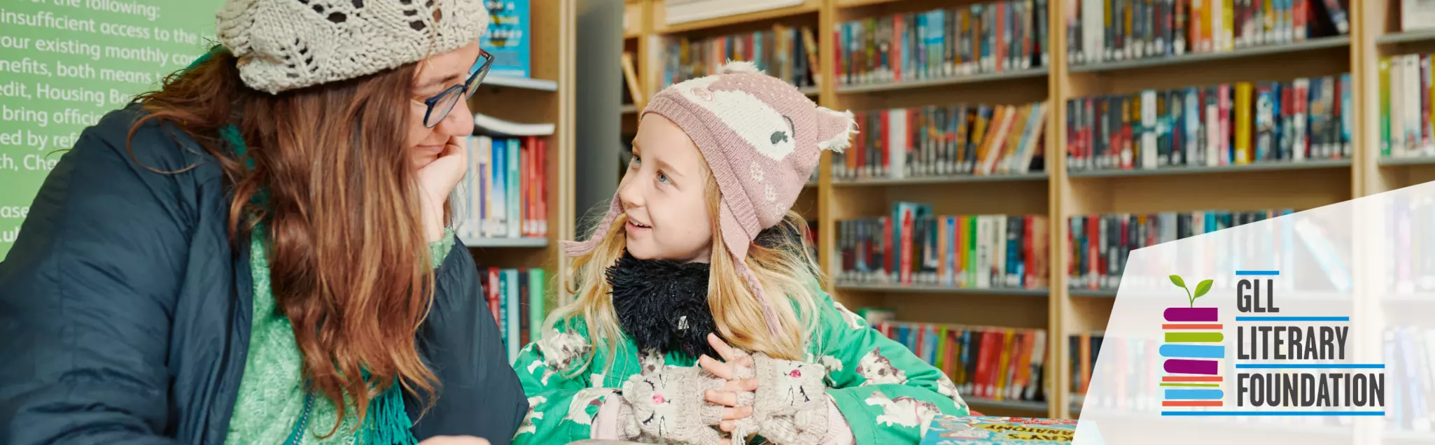 Adult and child in winter clothes smiling and reading together at a table in a library.