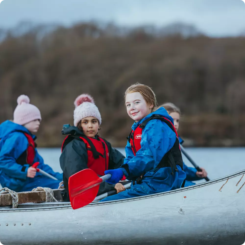 Children in waterproof gear paddling a canoe, with one girl smiling at the camera.