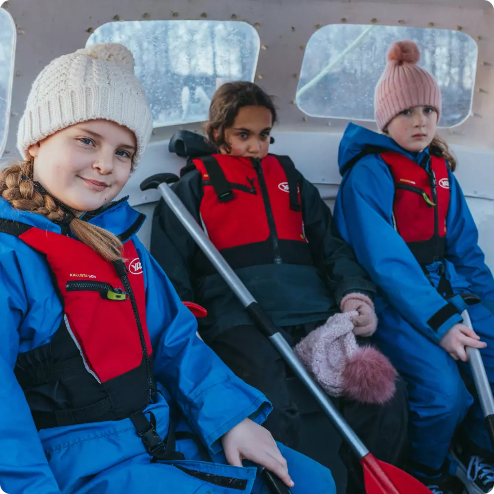 Three children in waterproof gear and life jackets sitting together in a boat.
