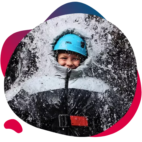 Smiling child in outdoor gear and helmet being splashed with water.