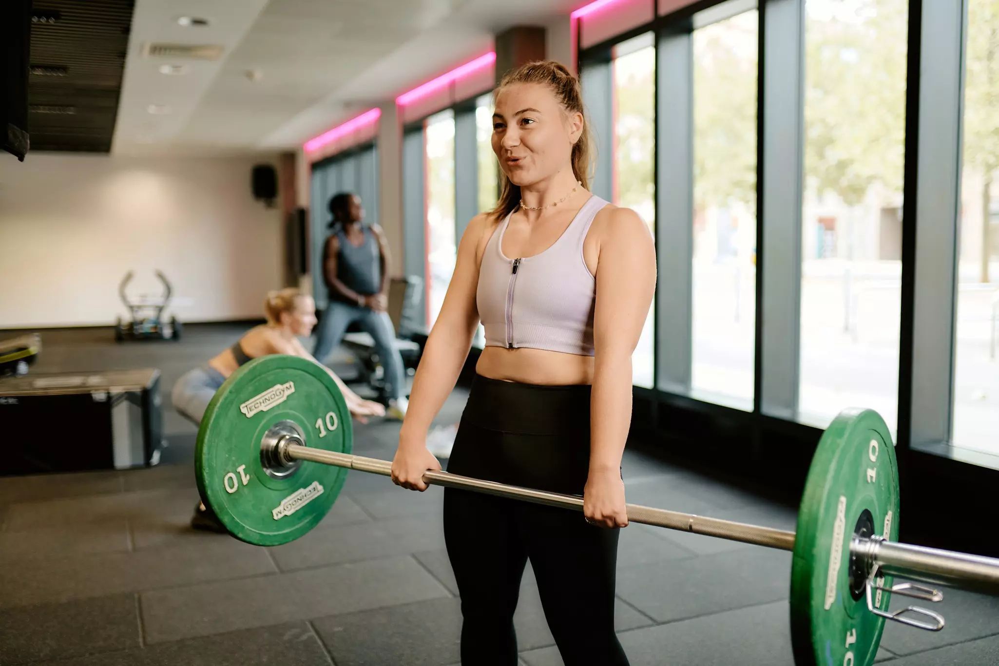 Woman lifting barbell with green plates in a gym with other members in background.