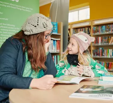 Adult and child in winter clothes smiling and reading together at a table in a library.