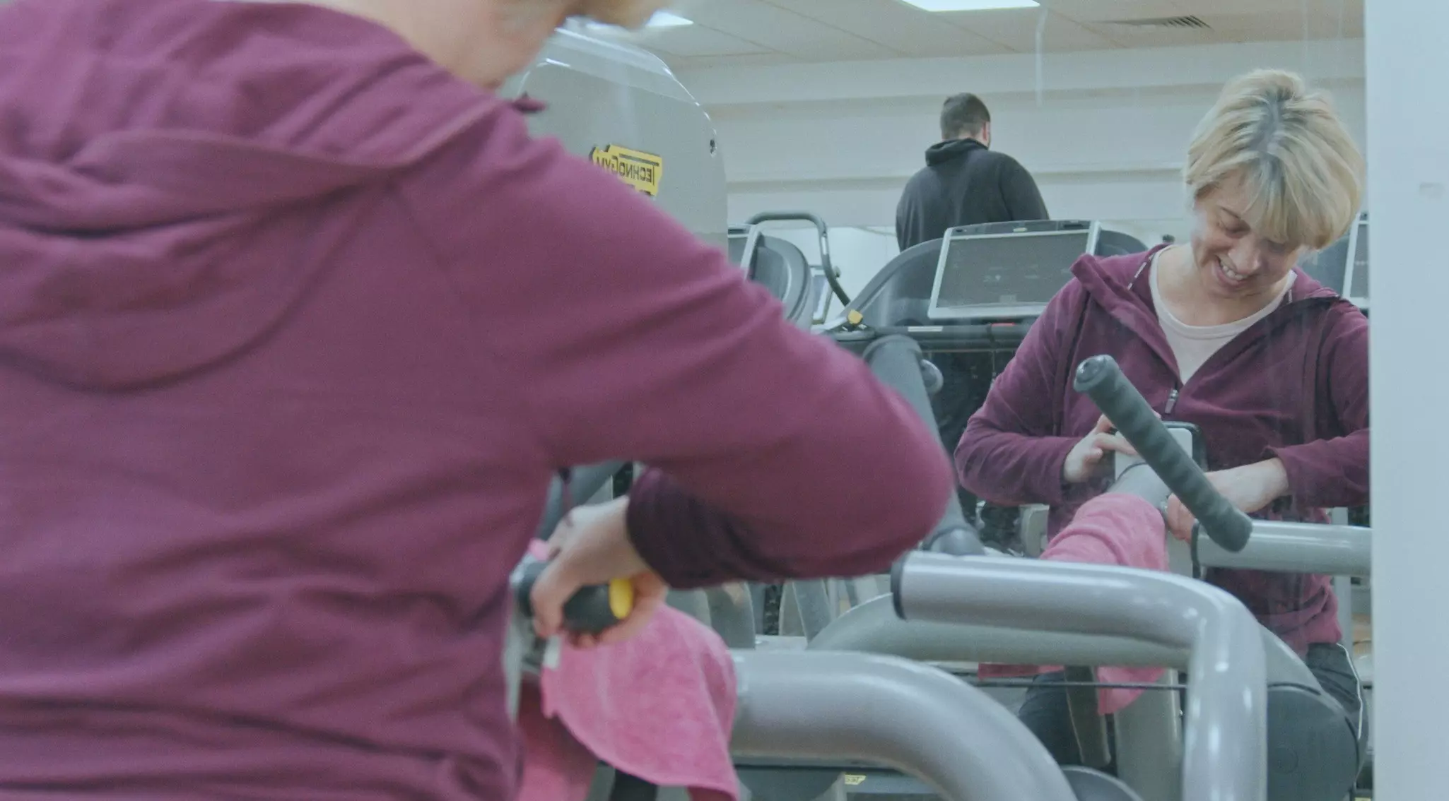 Woman in maroon jacket smiling in a gym with exercise equipment and other members in background.