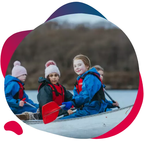 Children in waterproof gear paddling a canoe, with one girl smiling at the camera.