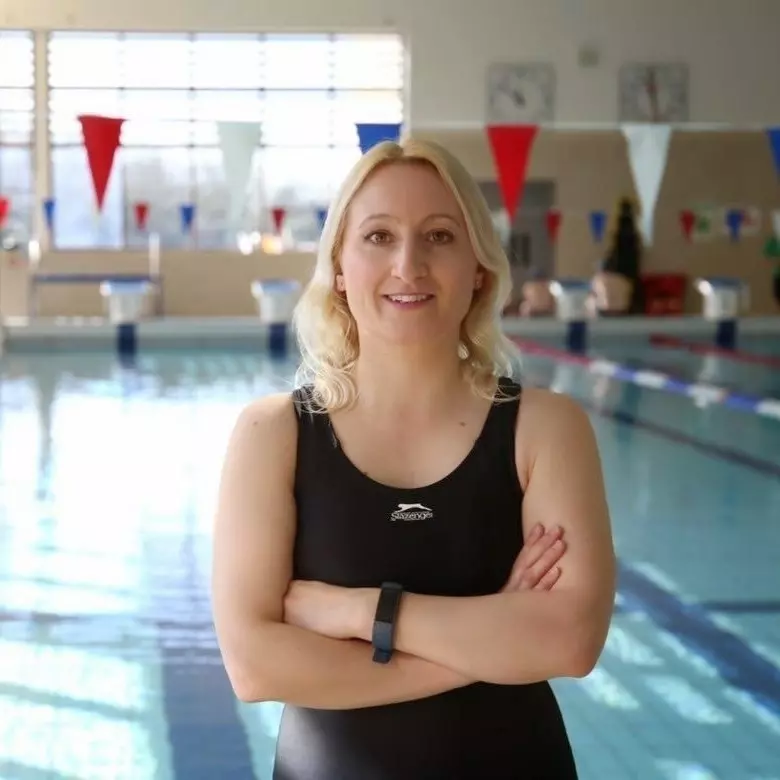 Woman with arms crossed wearing black sleeveless top by an indoor swimming pool.