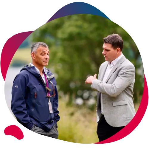 Two men talking outside in a green lakeside setting.
