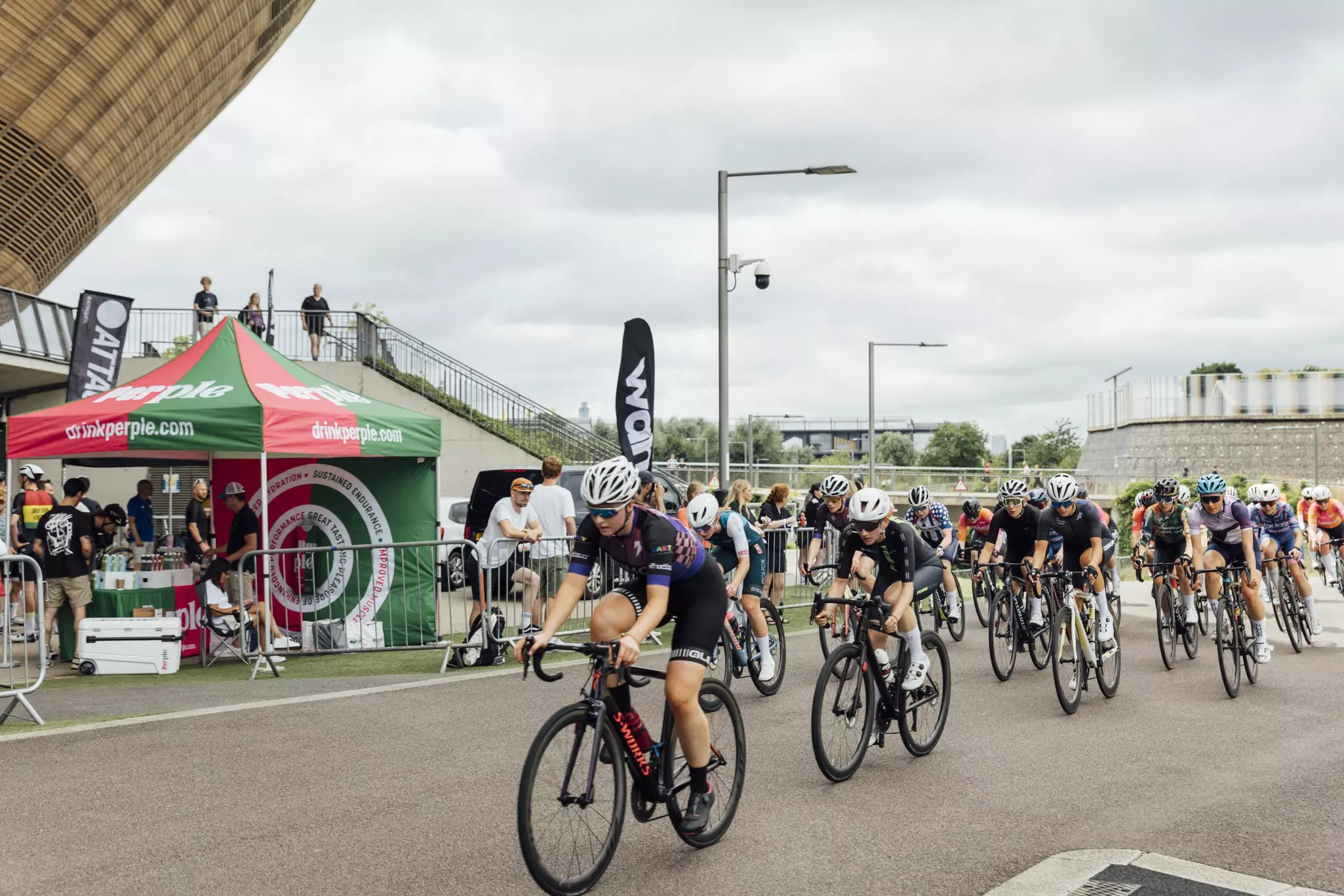Cyclists racing in a road cycling event near a velodrome, with spectators and sponsor tents nearby.