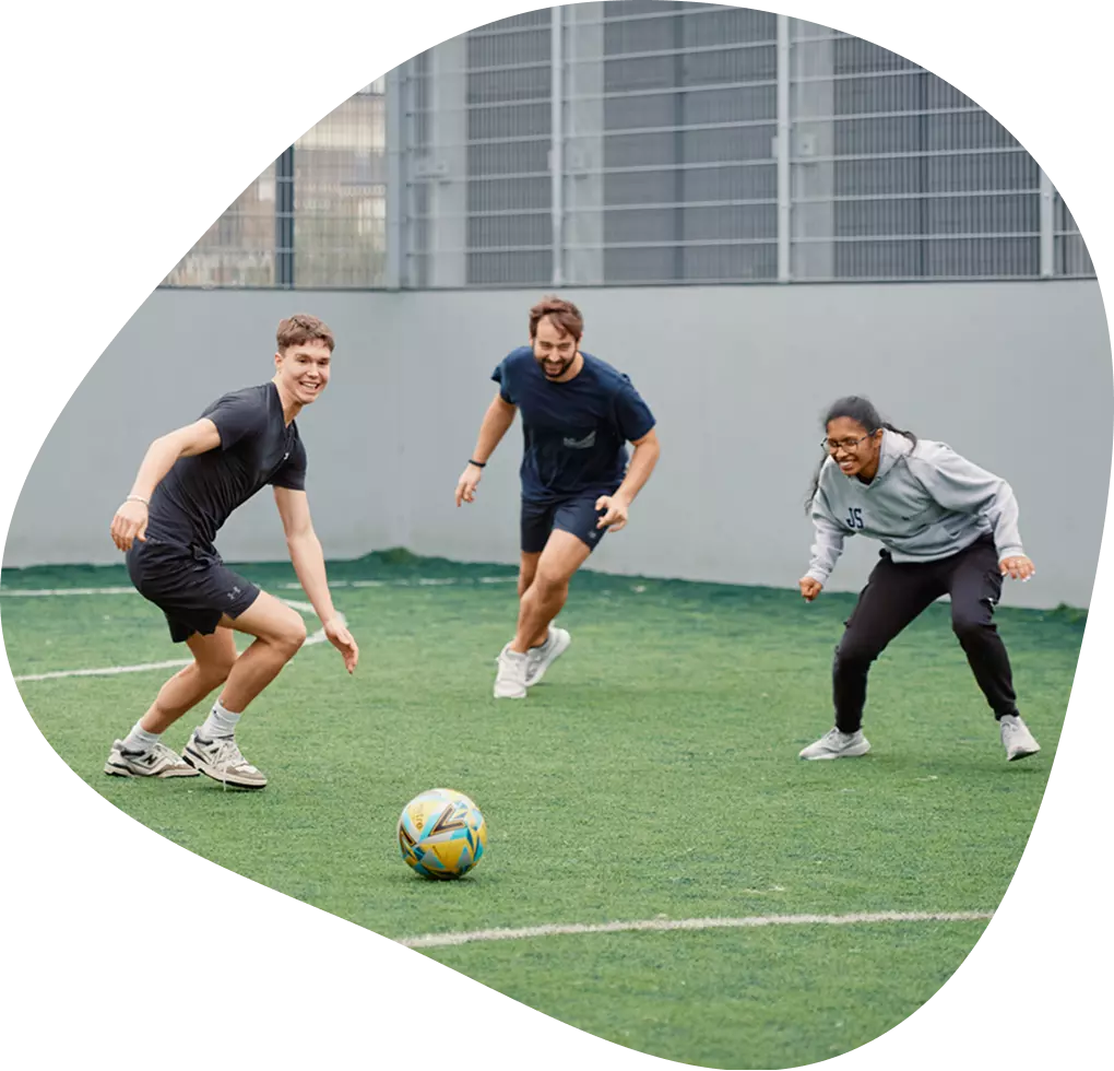 Three adults playing football on an outdoor pitch.