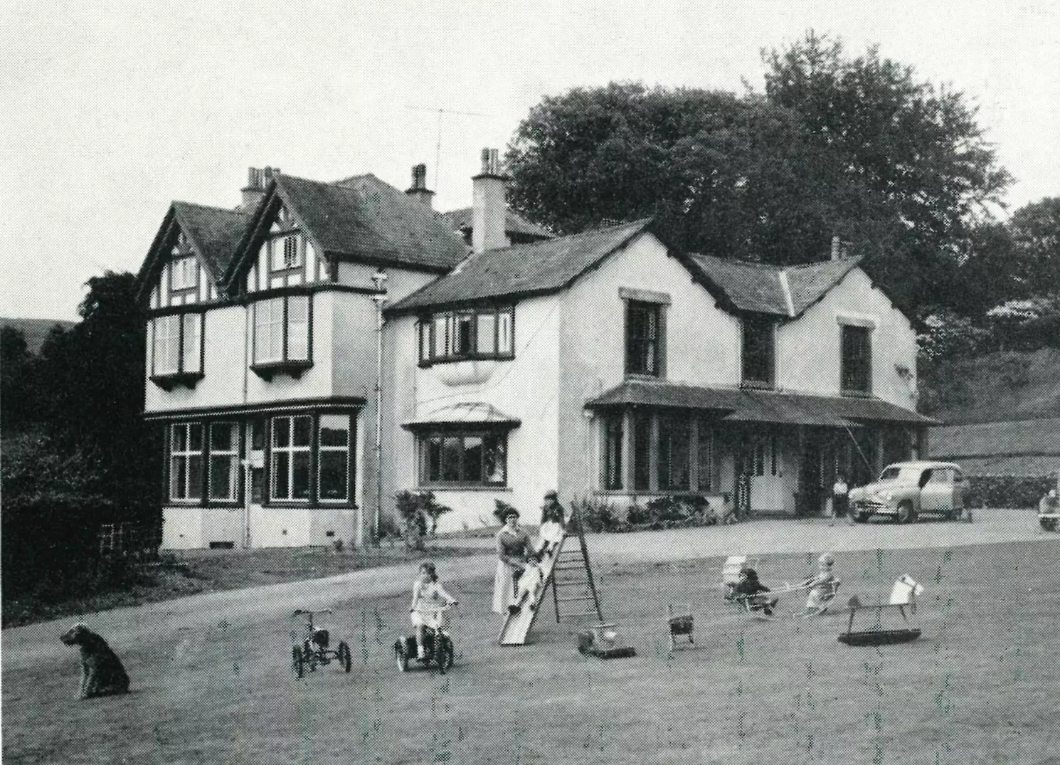 Vintage photo of a large house with children playing outside on tricycles and a slide.