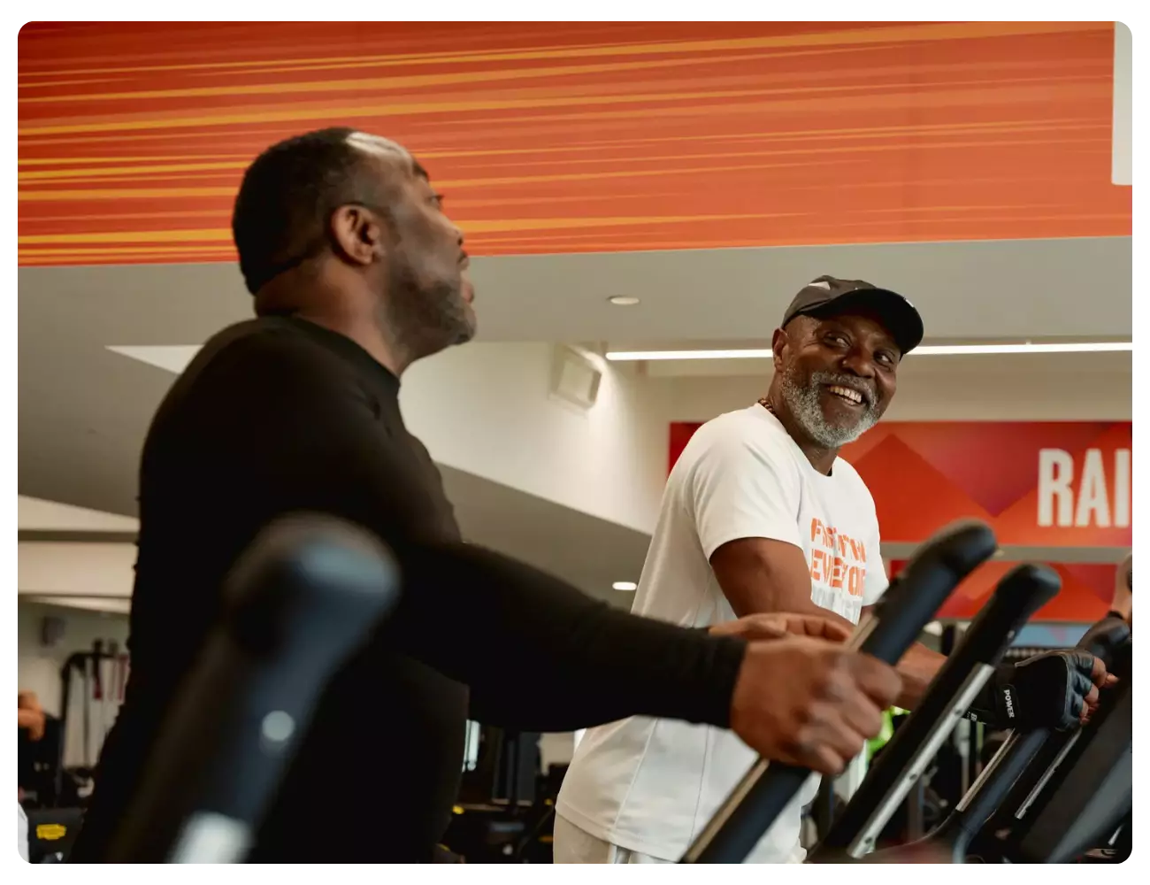 Two men smiling while exercising on cardio machines at the gym.