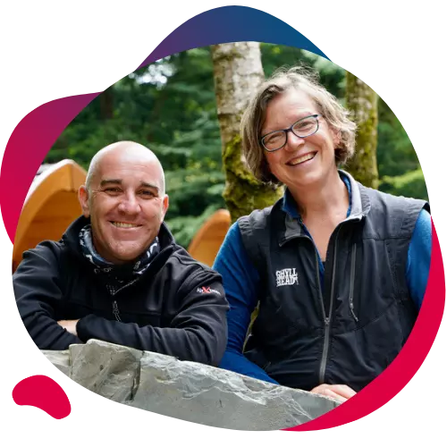 Two smiling people outdoors, leaning on a stone wall with trees in the background.