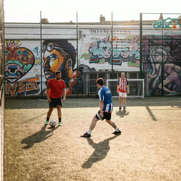Men playing football on an outdoor urban pitch with graffiti backdrop.