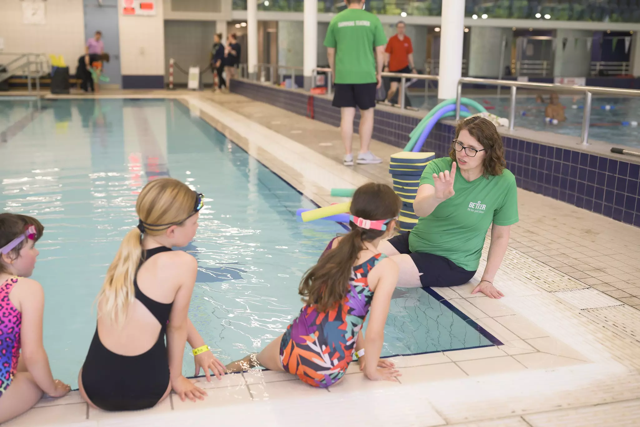 Swimming instructor sitting by the pool giving guidance to a group of young children in swimsuits.
