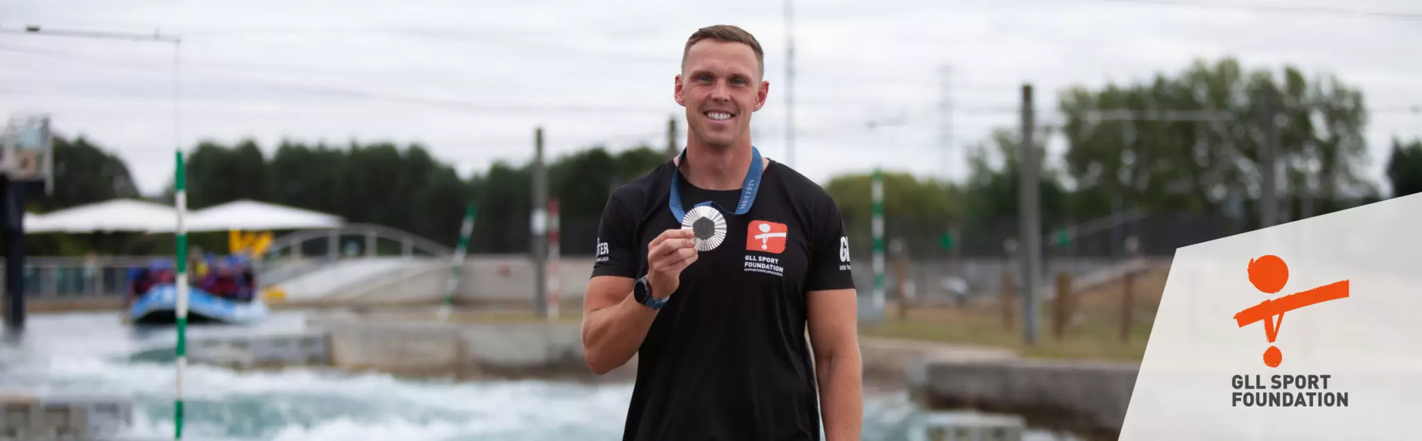 Athlete smiling and holding a medal at a water sports venue, wearing GLL Sport Foundation kit.