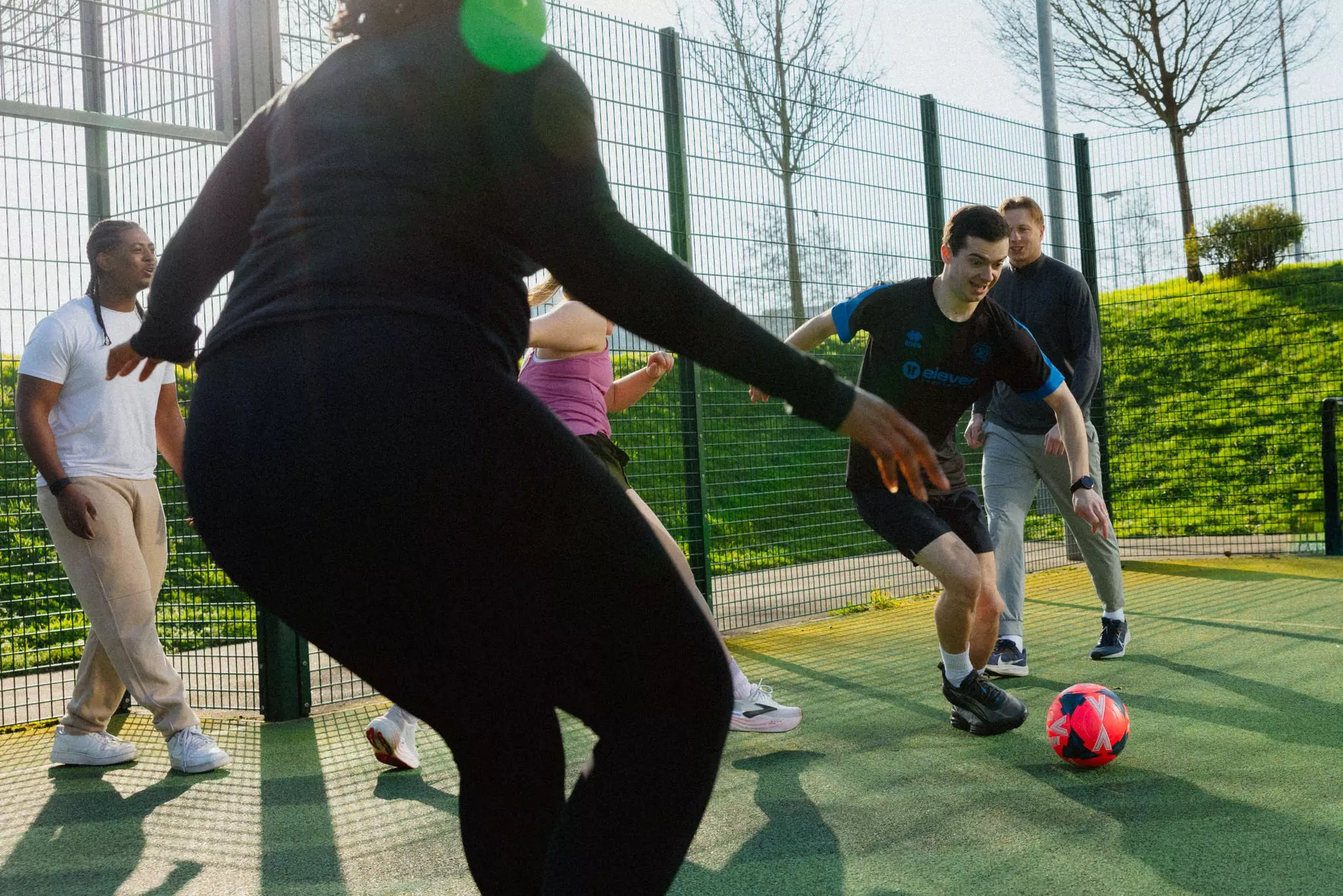 Group of people playing soccer on an outdoor court with green fencing and hedges.